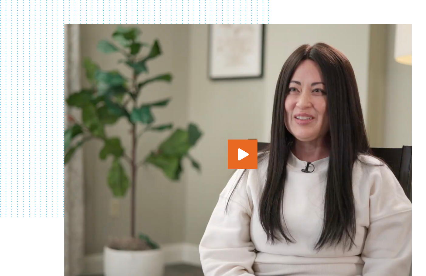 A smiling woman with long black hair wearing a white sweater, seated indoors with a potted plant and lamp in the background, and an orange play button icon overlaid on her shoulder, suggesting a video thumbnail.