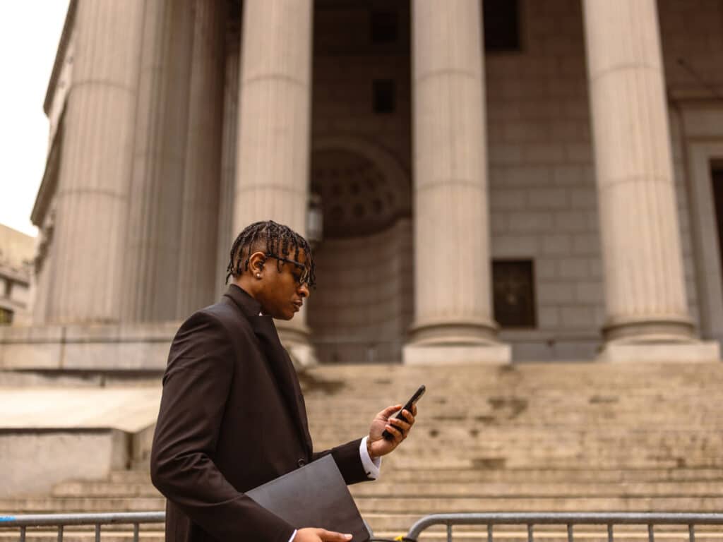 An African American businessman walks in the foreground, while looking at his phone and carrying a folio. In the background are classic roman columns and stone steps to a government building.